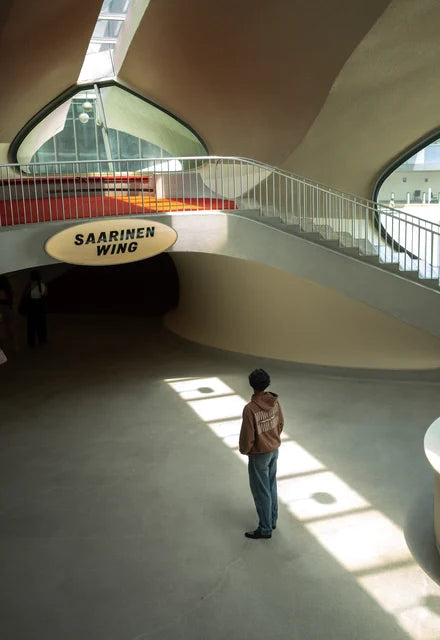 A traveller alone under the Saarinen wing sign in the TWA terminal at JFK, standing in a pane of sunlight